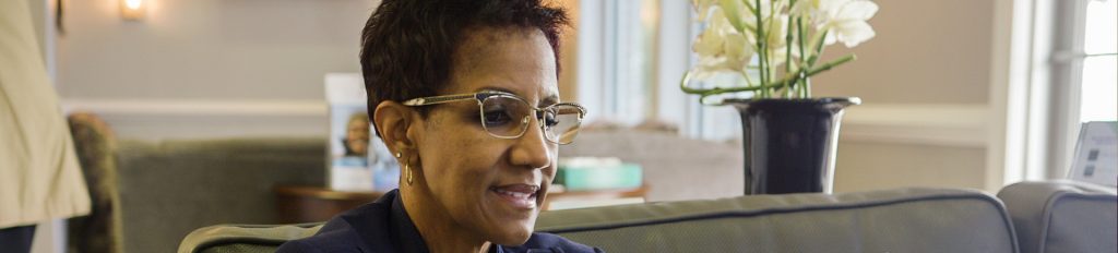A close-up profile of a woman with short dark hair and gold-rimmed glasses, wearing a navy blue blazer and gold hoop earrings